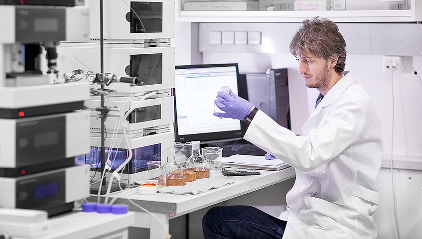 Das Bild zeigt eine Person im Labor mit Handschuhen, die ein reagenzglas hält. Im Raum befinden sich unterschiedliche Geräte und Computer.